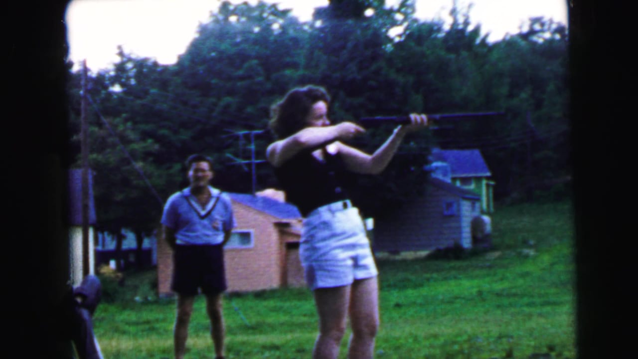 8mm home movie still, c.1964, possibly Camden NJ: young woman bracing a rifle in suburban backyard while male companion smiles nearby.