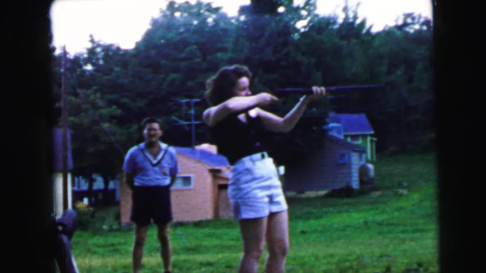 8mm home movie still, c.1964, possibly Camden NJ: young woman bracing a rifle in suburban backyard while male companion smiles nearby.