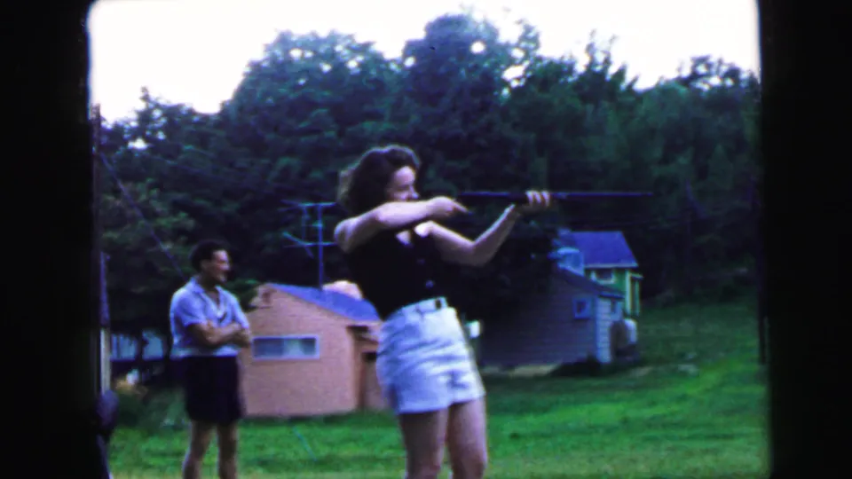 8mm home movie still, c.1964, possibly Camden NJ: young woman completing a rifle shot while male companion reacts nearby in a grassy backyard.