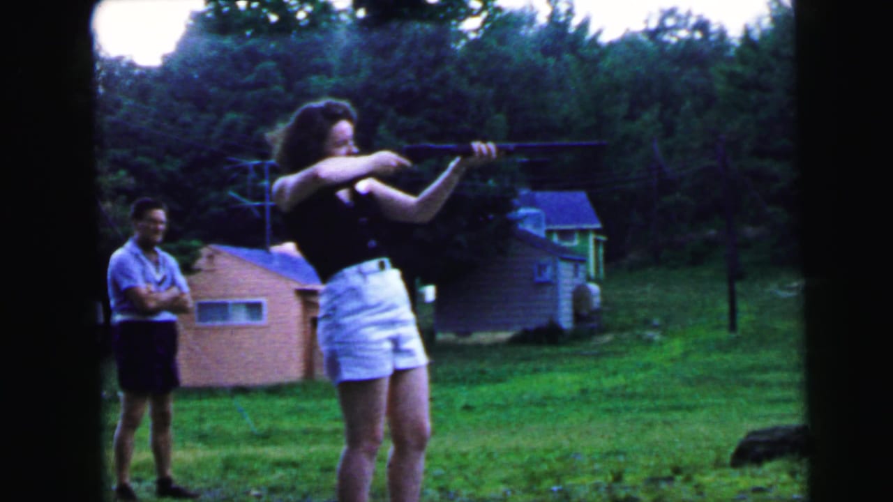 Woman aiming a long gun in a 1964 Camden, New Jersey suburban backyard on 8mm film; pink house and observing man in light blue shirt visible behind her.