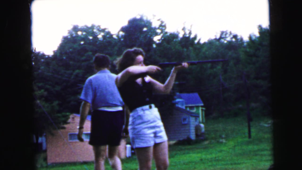 Woman and man seen from behind; she holds a long gun extended to the right, he stands close in a light blue shirt, with pink and blue houses in the background.