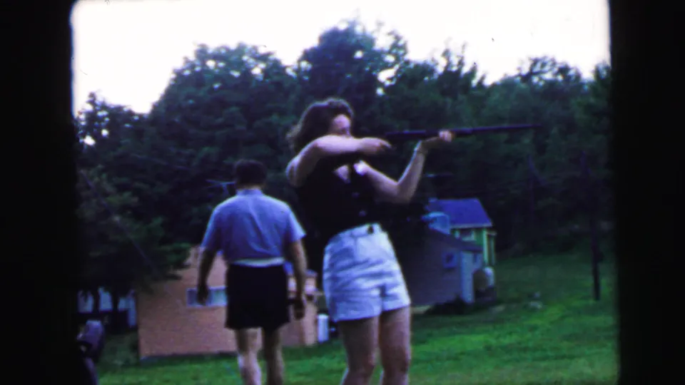 Woman holding long gun with male companion at her left side in a grassy backyard, circa 1964, Camden, New Jersey, 8mm home movie.