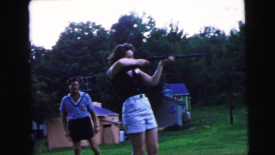Woman lowering long gun as man turns toward camera in 1964 suburban backyard, Camden, New Jersey; man appears to react or smile, 8mm film.