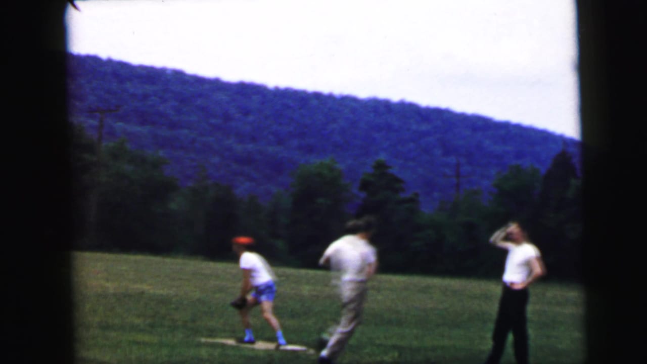 Group of men playing an outdoor game on a grass field circa 1964, attributed to Camden, New Jersey; small orange disc in flight; forested ridge and overcast sky behind.