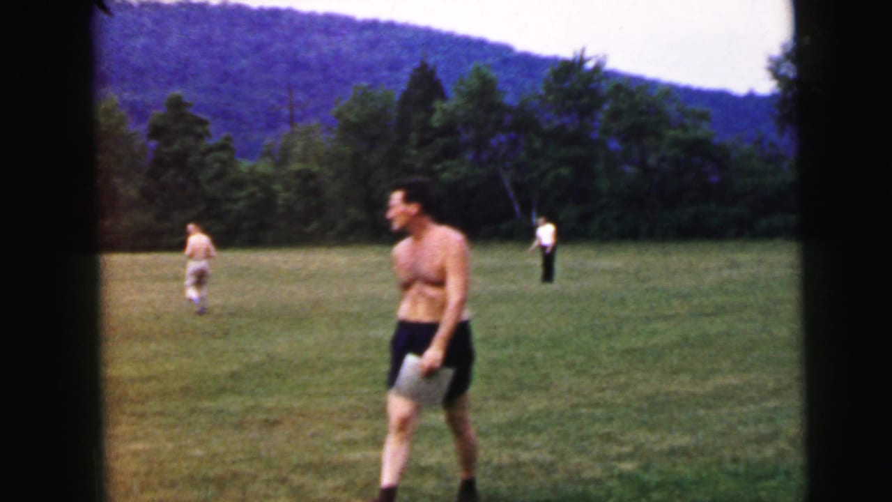 Shirtless man carrying white papers or sheets stands on an open grass field circa 1964, attributed to Camden, New Jersey; utility pole and forested ridge visible behind.