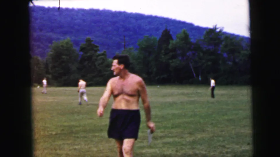 Shirtless man smiling and walking toward camera on an open summer grass field circa 1964, attributed to Camden, New Jersey; scattered figures and forested ridge visible in background.