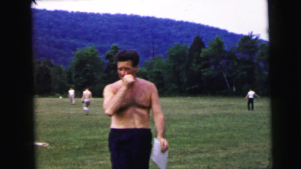 Shirtless man holding white papers and raising hand to mouth, standing on a summer grass field circa 1964, attributed to Camden, New Jersey; forested hillside and background players visible.