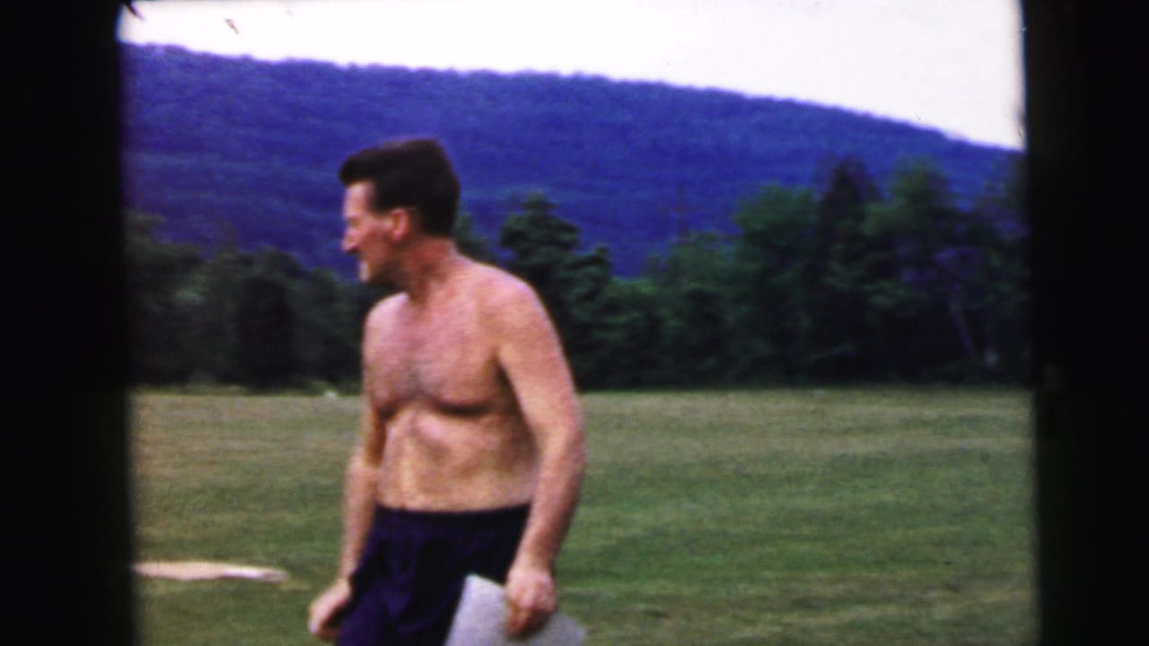 Shirtless dark-haired man walking across a grassy field with wooded rolling hills in the background, 8mm home movie.