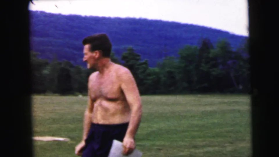 Shirtless man in dark trousers walks across an open field; forested hills rise behind him in what metadata identifies as a 1964 home movie.