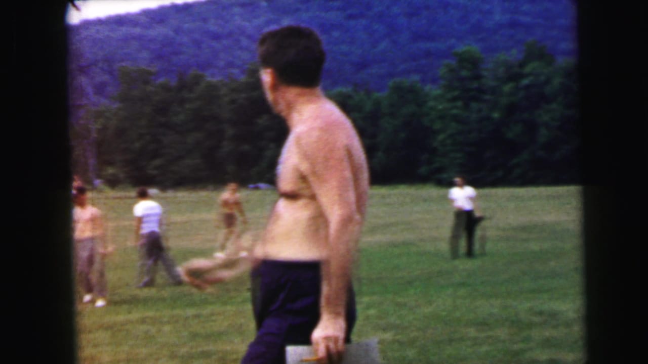 Shirtless man turns mid-stride on an open field; background shows multiple casually dressed figures and a forested hillside, 8mm home movie circa 1964.