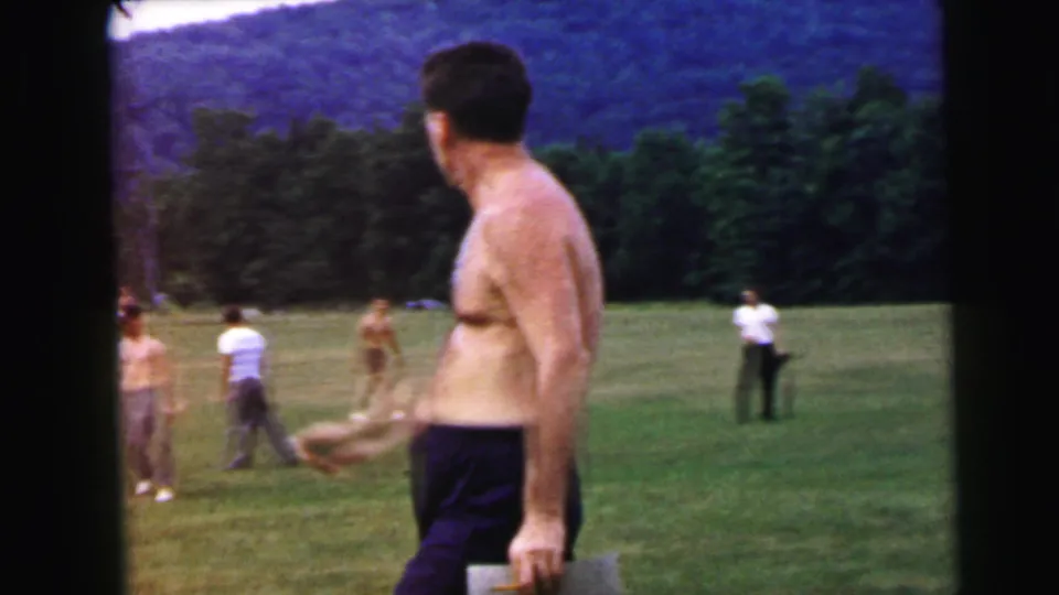 Shirtless man turns mid-stride on an open field; background shows multiple casually dressed figures and a forested hillside, 8mm home movie circa 1964.