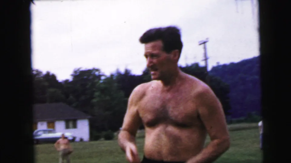 Shirtless man approaches camera smiling; behind him stand a white mid-century house, a parked car, utility poles, and trees, 8mm home movie circa 1964.