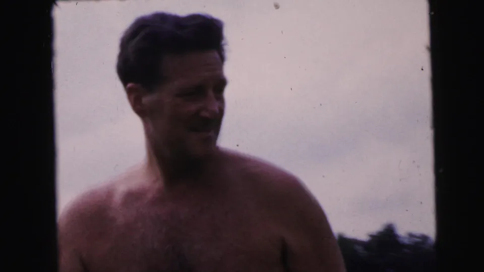 Tight close-up of shirtless dark-haired man looking toward the camera; background washed to bright sky, 8mm home movie circa 1964.