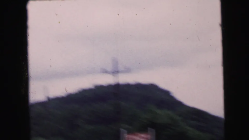 8mm frame circa 1964 from a moving vehicle: wooded ridgeline under overcast sky, a faint utility or transmission tower on the hilltop, and a partial structure at the lower frame edge.