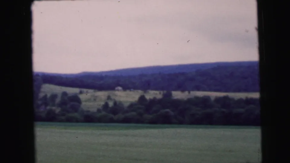 8mm home movie frame circa 1964 — car window view of open rural fields and a tree-lined ridgeline, attributed to the eastern United States.