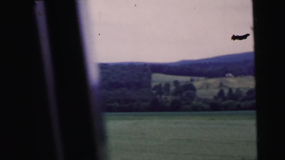 8mm home movie frame circa 1964 — car window POV over rolling rural landscape with wooded ridges; a small dark object in sky may be a bird or film artifact.