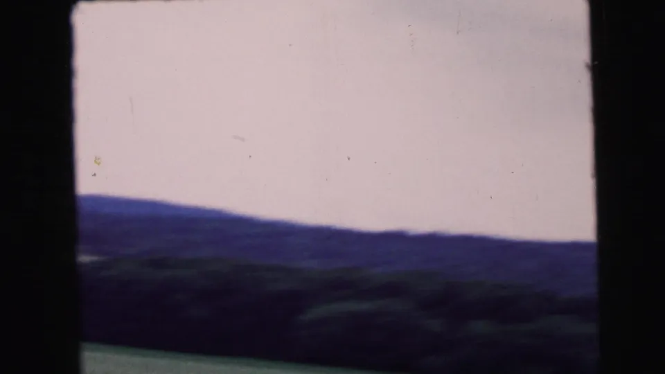 8mm home movie frame circa 1964 — open rural landscape with broad overcast sky and low tree-lined horizon, shot from a moving vehicle.