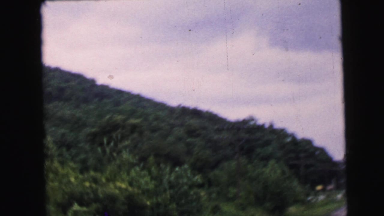 8mm home movie shot from a moving vehicle, showing dense deciduous hillside and overcast sky, reportedly filmed in New Jersey in 1964.