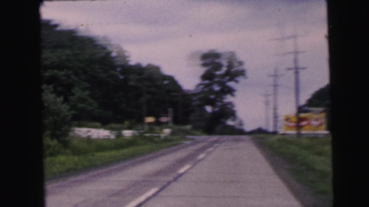 8mm home movie forward POV on a two-lane American road, circa 1964, with roadside signs, grass verge, trees, and high-tension transmission pylons in the background.