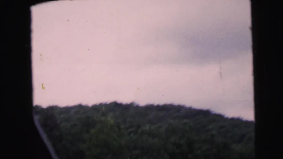 8mm home movie frame dominated by an overcast sky with tree-covered hills at the lower edge, filmed from a moving vehicle, reportedly New Jersey 1964.