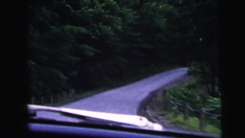 8mm home movie windshield POV of a curving two-lane road bordered by dense summer foliage, car hood visible in the lower frame, reportedly New Jersey 1964.