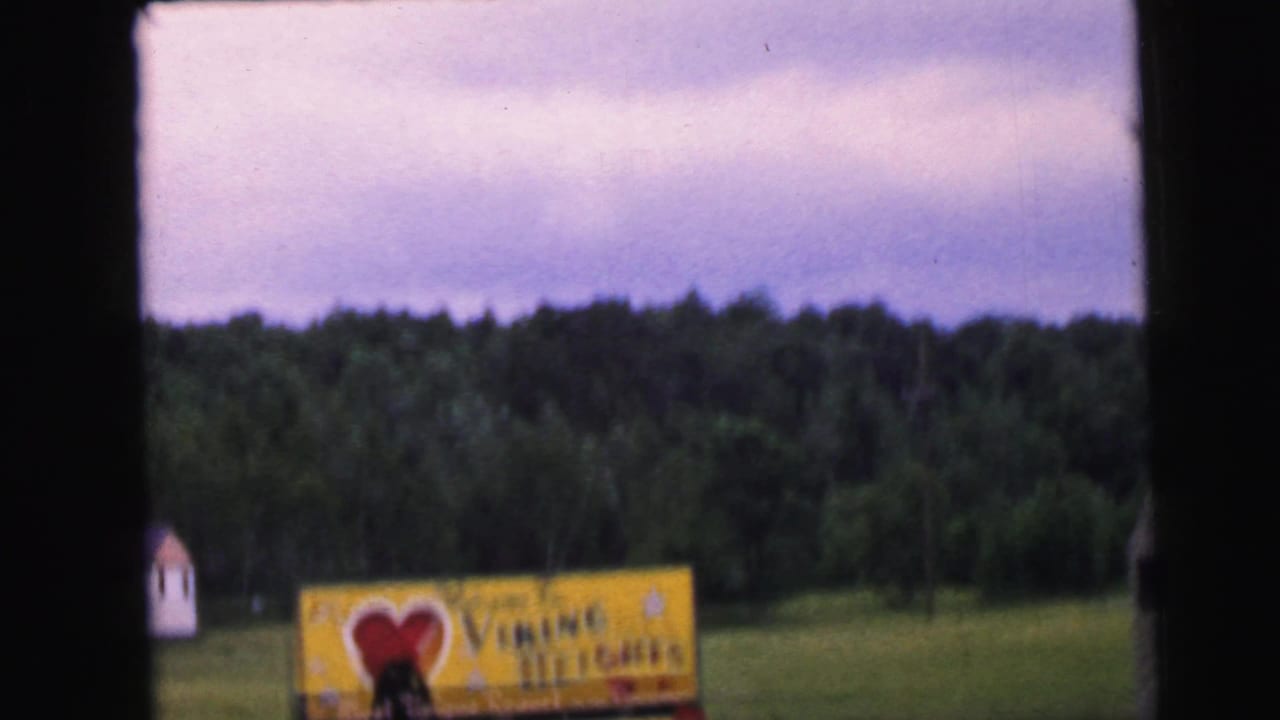 8mm home movie frame circa 1964 showing the Viking Heights welcome sign at a distance, approached by car, with a small white outbuilding at left and a dense treeline behind.