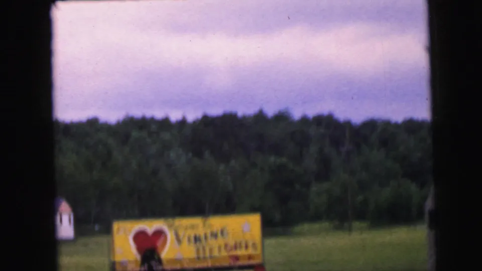 8mm home movie frame circa 1964 showing the Viking Heights welcome sign at a distance, approached by car, with a small white outbuilding at left and a dense treeline behind.