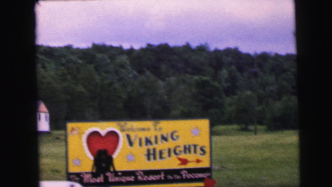 8mm home movie frame circa 1964 showing the Viking Heights resort welcome sign clearly, with red heart, star motifs, a red arrow, tagline text at the base, and a silhouetted figure nearby.