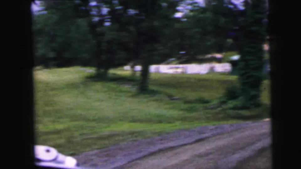 8mm home movie frame circa 1964 after passing the Viking Heights sign; green lawn, a gravel road edge, scattered deciduous trees, and low background structures suggest the resort entrance area.