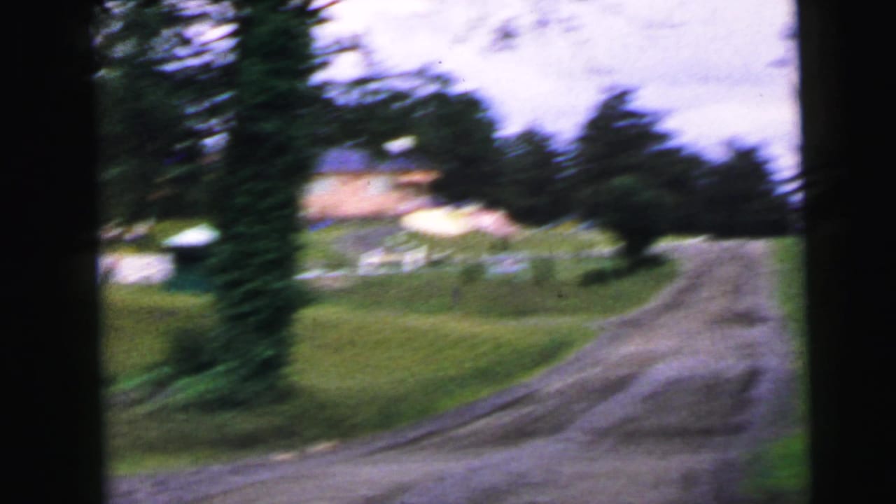 8mm home movie frame circa 1964 showing a dirt access road with an evergreen tree at left, grassy verges, and distant structures in what may be the Camden, New Jersey area.