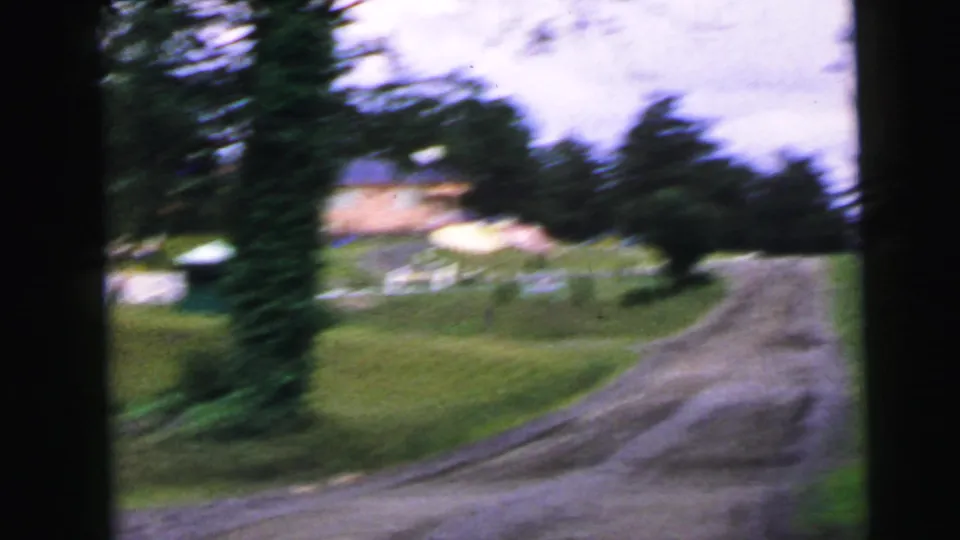 8mm home movie frame circa 1964 showing a dirt access road with an evergreen tree at left, grassy verges, and distant structures in what may be the Camden, New Jersey area.