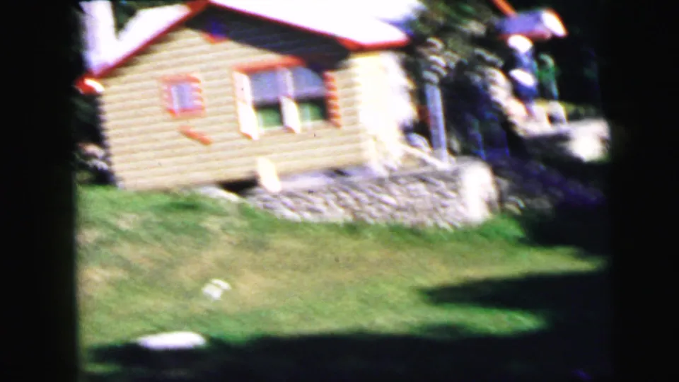 8mm frame circa 1964 showing a small log cabin with red-trimmed windows and green lawn, possibly a resort cabin at Viking Heights, New Jersey; partial human figure visible at right edge.