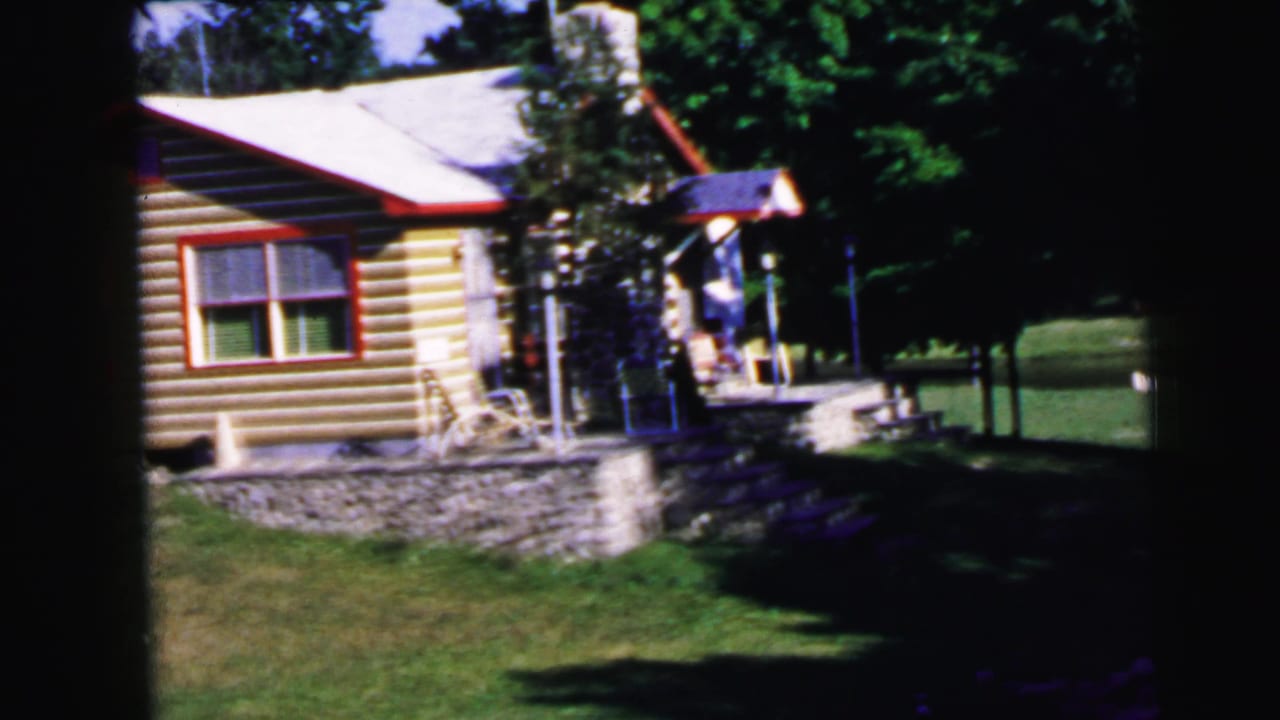 8mm home movie frame, c.1964: log cabin with stone rubble foundation, red window trim, and a person near the porch in a wooded summer setting.
