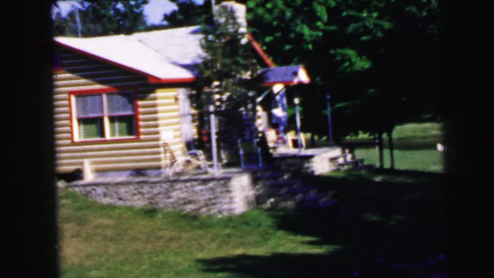 8mm home movie frame, c.1964: log cabin with stone rubble foundation, red window trim, and a person near the porch in a wooded summer setting.