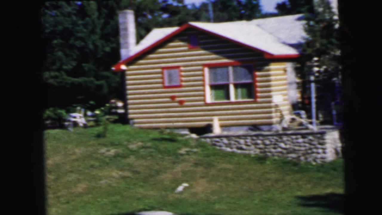 8mm film frame, c.1964: log cabin exterior showing red window trim, rubble stone foundation, roofline with chimney, surrounded by green summer foliage.