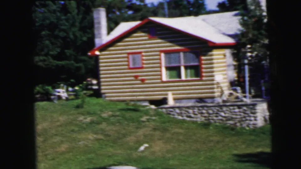 8mm film frame, c.1964: log cabin exterior showing red window trim, rubble stone foundation, roofline with chimney, surrounded by green summer foliage.