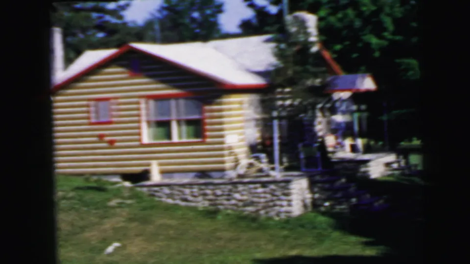 8mm film frame, c.1964: log cabin with yellow-tan log walls, red window trim, stone foundation, and outdoor seating near the porch entry.