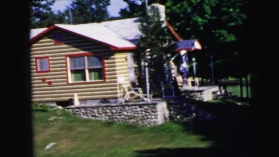8mm film frame, c.1964: log cabin with red-trimmed windows and stone foundation; a person is visible near the covered porch entry alongside outdoor chairs.