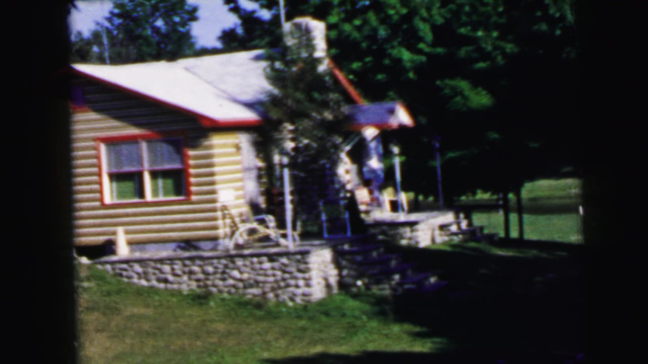 Log cabin with red-trimmed windows and stone retaining wall, dense trees behind, 8mm footage.