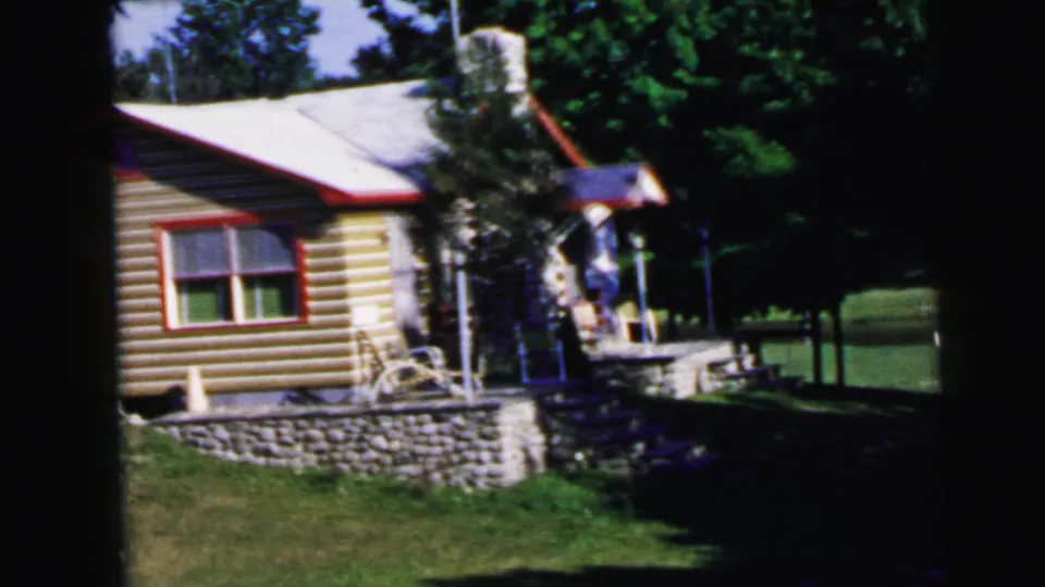 Mid-1960s log-sided cabin exterior with red window trim and stone retaining wall, home movie footage, United States.