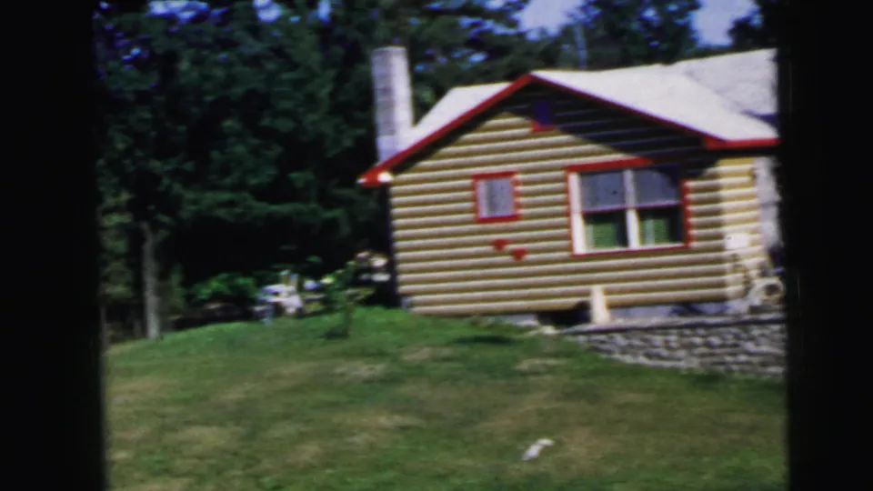 1960s log-sided cabin with cylindrical chimney, red trim, and stone retaining wall, home movie exterior shot, United States.