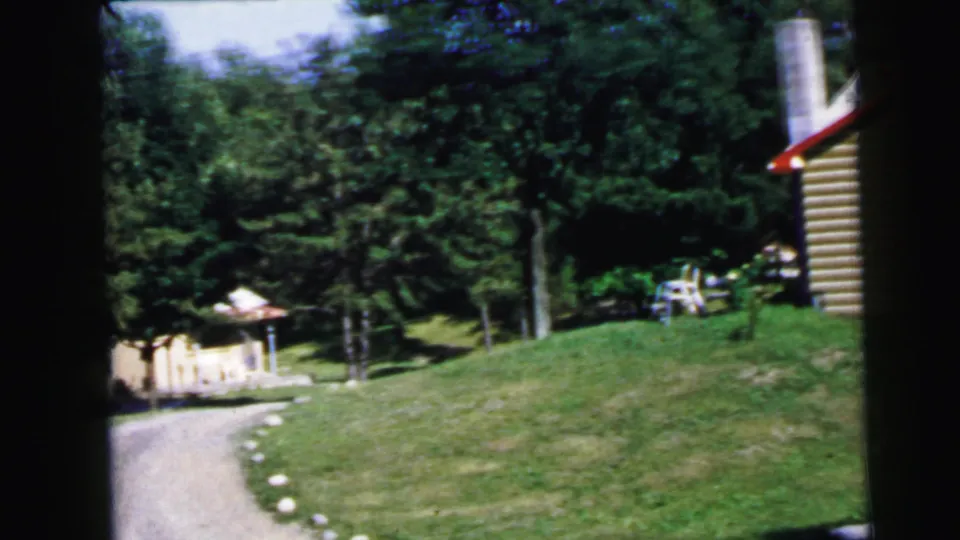 Wide 1960s home movie shot of wooded retreat property with curved gravel driveway, cylindrical structure, and small outbuildings, United States.