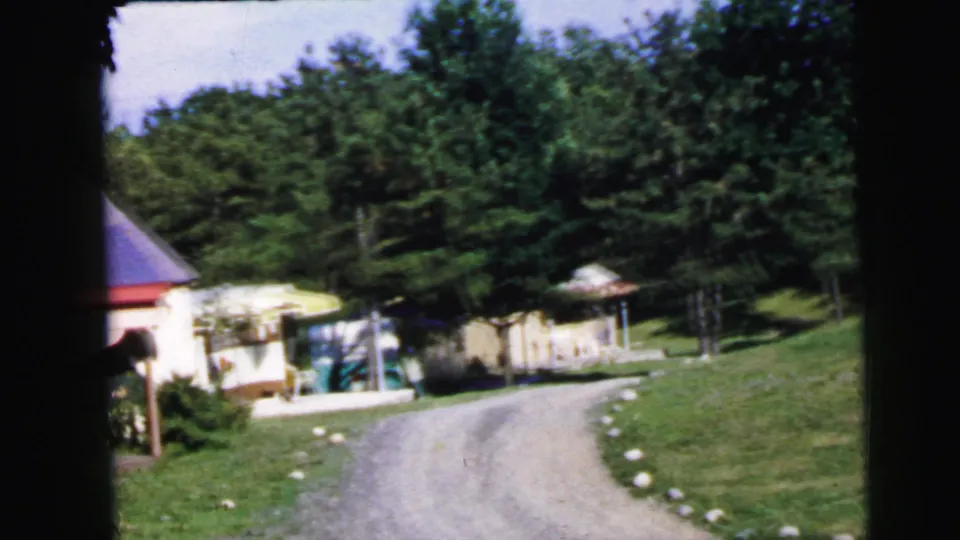 1960s home movie wide shot of camp or retreat compound with conical-roofed outbuilding, stone-bordered driveway, and dense mixed woodland, United States.