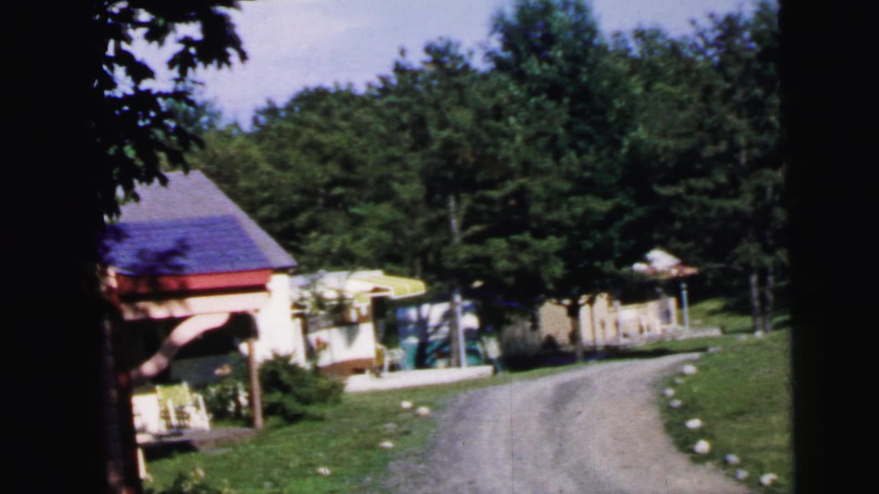 Gravel driveway curving past open pavilion with purple roof and colorful cabins, framed by summer trees, 8mm film.
