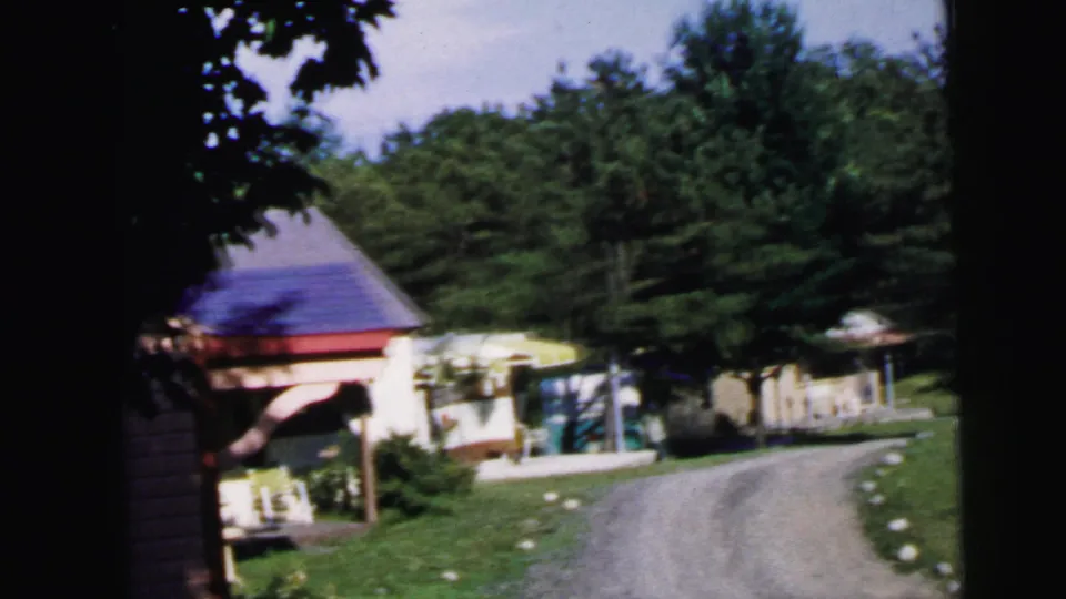 8mm frame from the same 1964 sequence; camera has advanced slightly along the gravel driveway toward the purple-roofed pavilion and adjacent cabin structures.