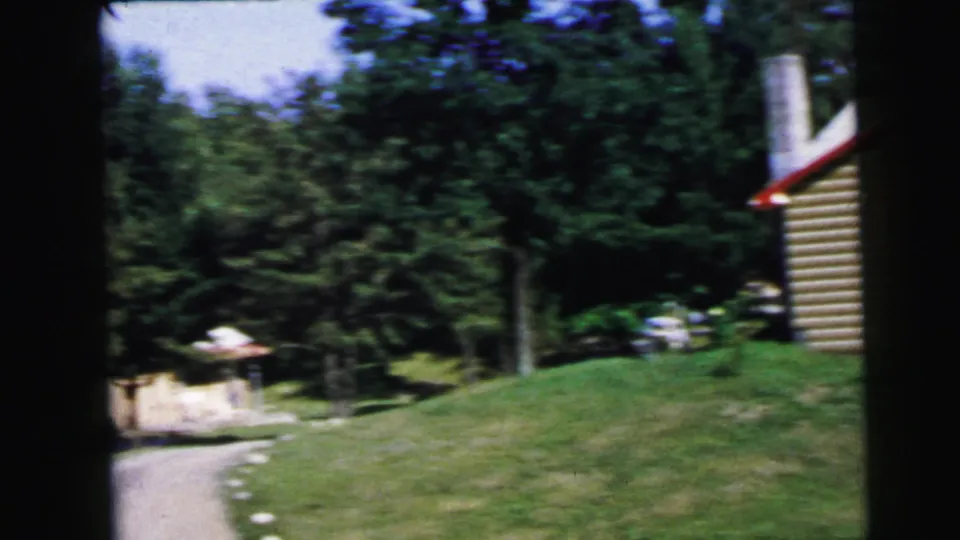 8mm frame from 1964 showing a tan log-sided cabin with red trim and a white roof; a cylindrical silo-like structure is visible at left among coniferous and deciduous trees.
