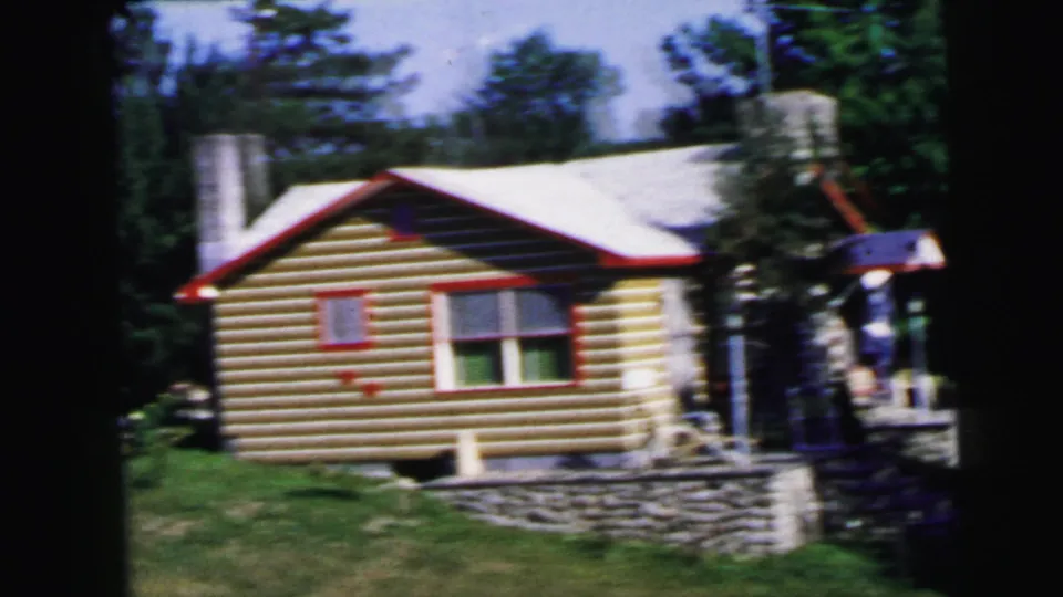 8mm frame from 1964 with camera closer to the log cabin, showing red-framed windows, a stone or concrete block foundation, and outdoor equipment to the right under a bright summer sky.