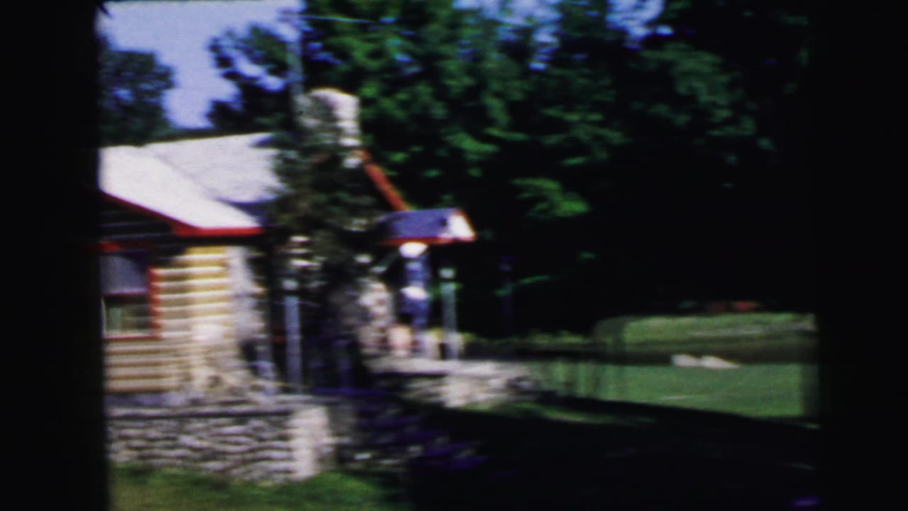 Log-sided house with stone foundation wall and red trim, surrounded by green trees, seen from the yard.