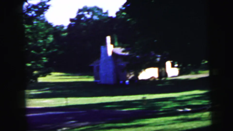 8mm home movie frame, c. 1964, showing a small building with a white chimney and a dark vehicle parked nearby, set within a green suburban yard.