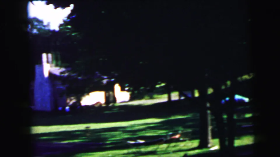 Underexposed 8mm home movie frame of a heavily shaded yard in summer, with tree trunks, patchy sunlight on lawn, and a white structure at the frame edge; consistent with Camden, New Jersey, 1964.